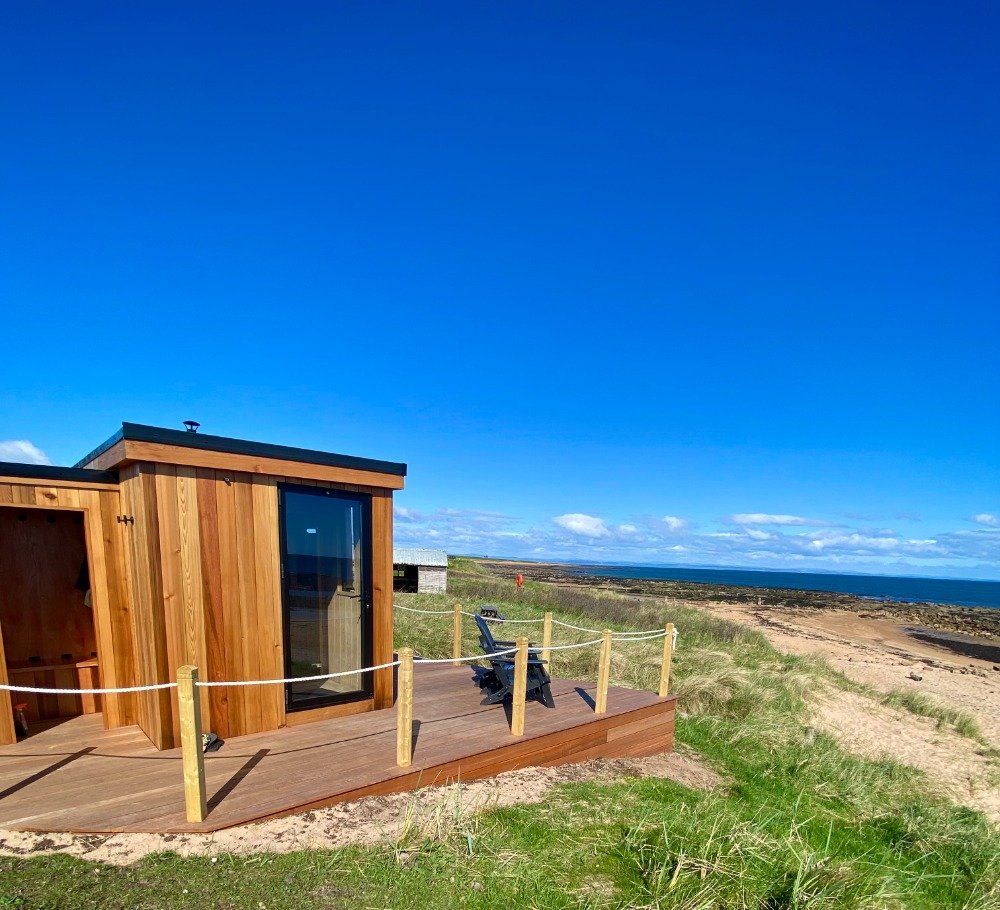 Wood-fired sauna on raised decking in sand dunes with chairs overlooking the beach and North Sea