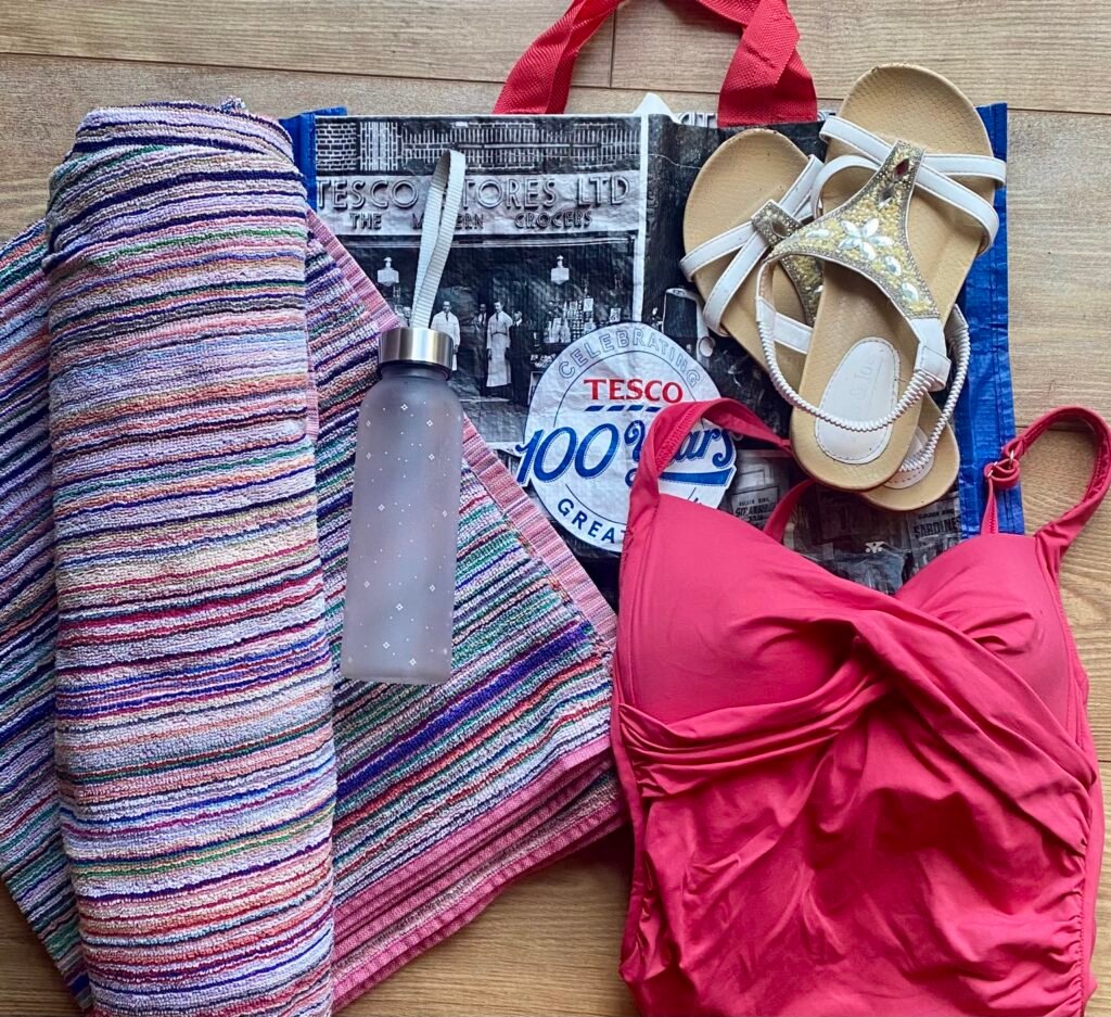 Beginner wild sauna essentials laid out beside a Tesco bag including towels, sandals, swimwear and water bottle
