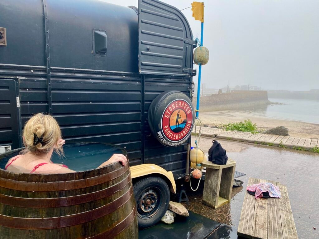 A person sitting in the whisky barrel cold plunge at Driftwood Sauna, looking out to sea