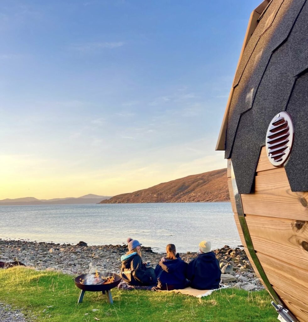 Applecross Sauna Wester Ross three people in dryrobes beside a fire pit on the shore at sunset next to the barrel sauna