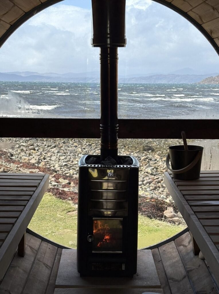 Applecross Sauna Wester Ross Harvia wood stove with a lit fire between slatted benches and a round window onto a choppy sea