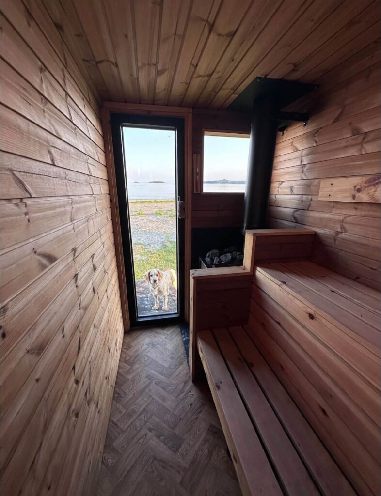 Bothan Jura Wild Sauna Isle of Jura timber sauna interior with tiered benches, wood stove and a view through the glass door to the sea