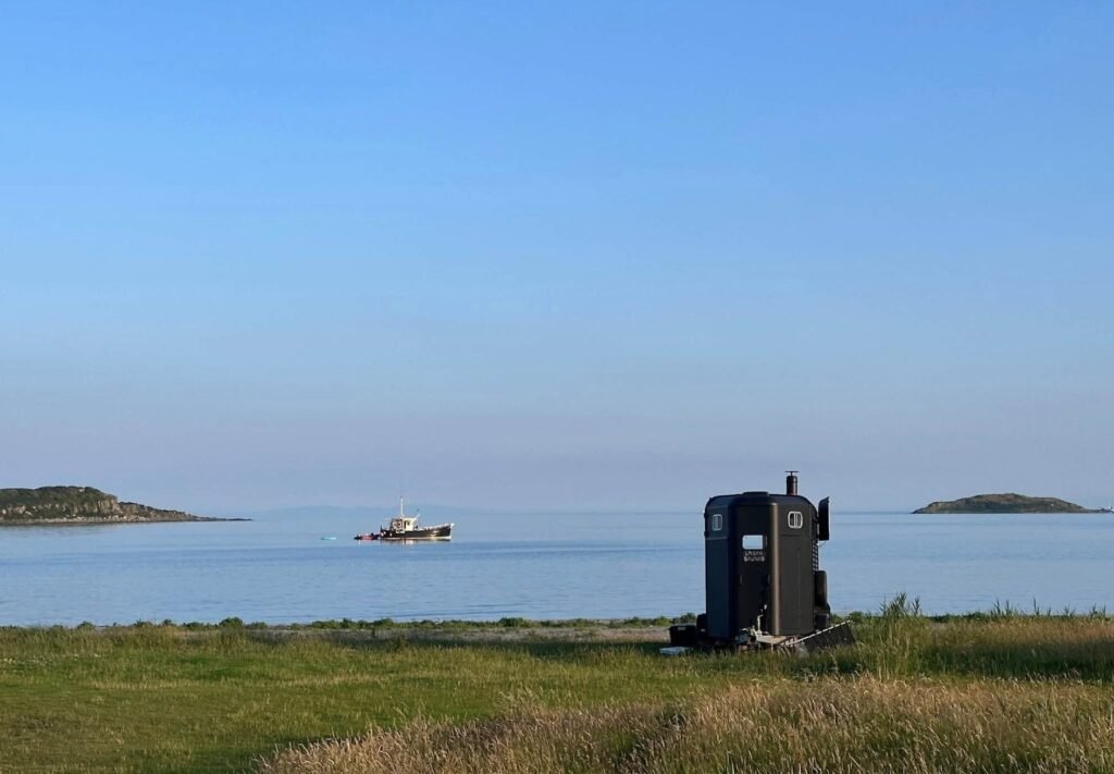 Bothan Jura Wild Sauna Isle of Jura horsebox sauna on the shore with a boat anchored in calm water and small islands beyond