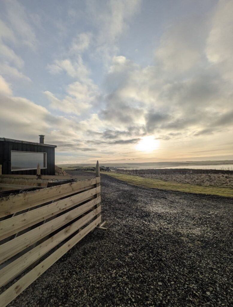 Seaview Sauna Dunnet Bay sauna cabin beside the timber-fenced plunge terrace with the bay and low sun beyond